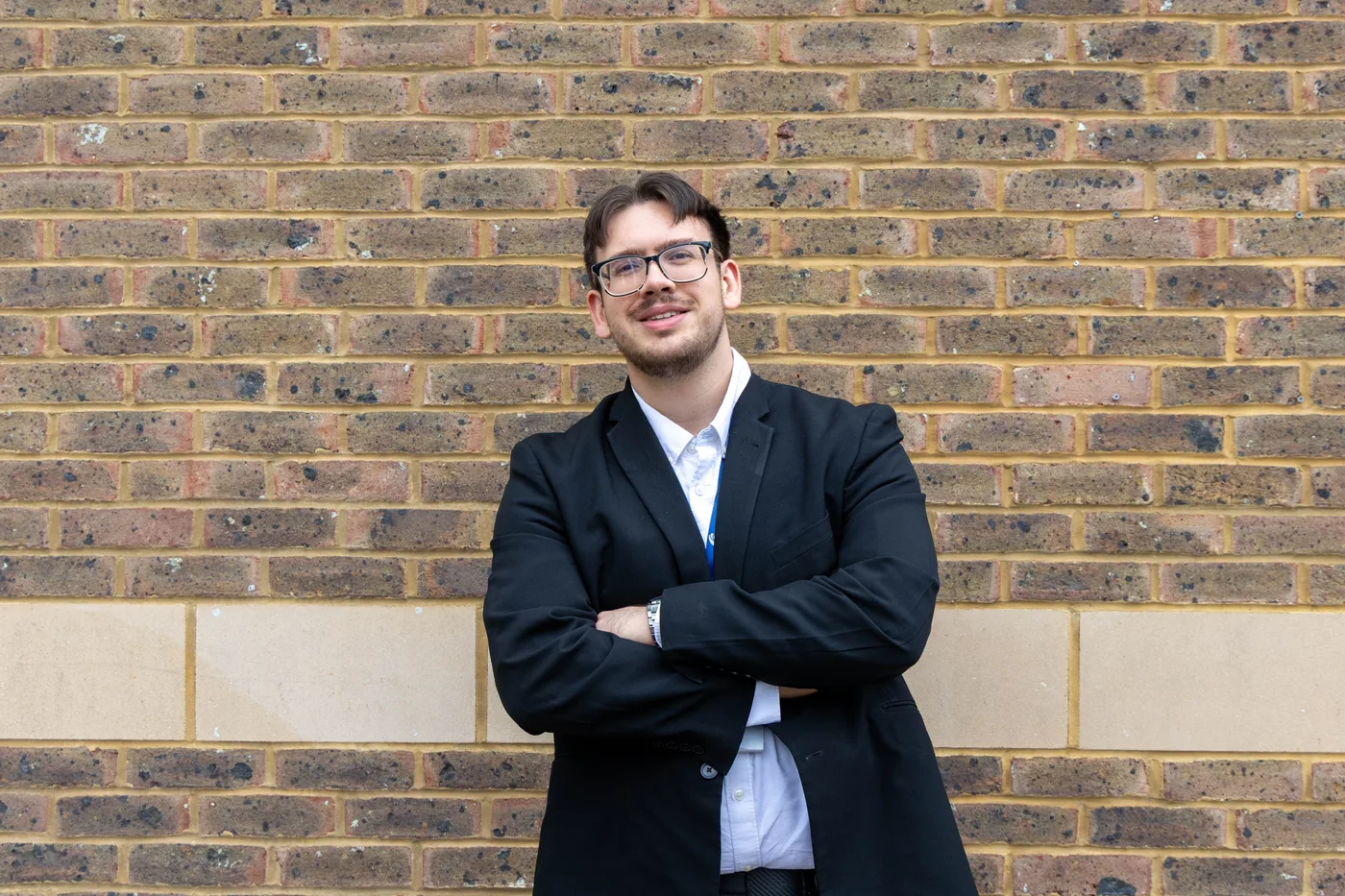 Alex Crean, in glasses, a dark jacket and white shirt, standing with his arms folded against a brick wall