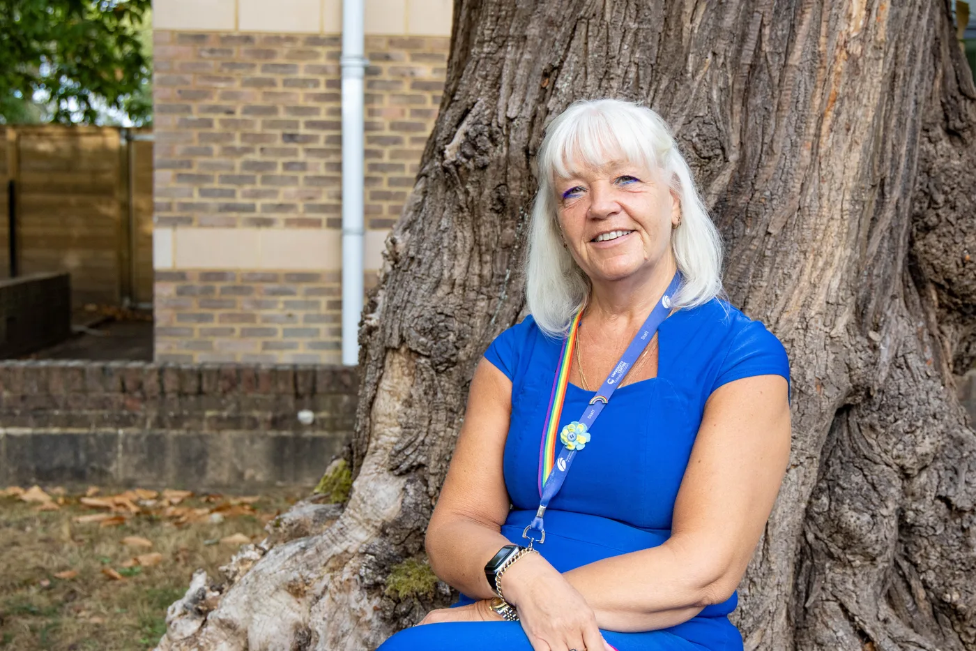 Michelle Chapman, in a blue dress and lanyard, sitting in front of a large tree trunk outside a brick building