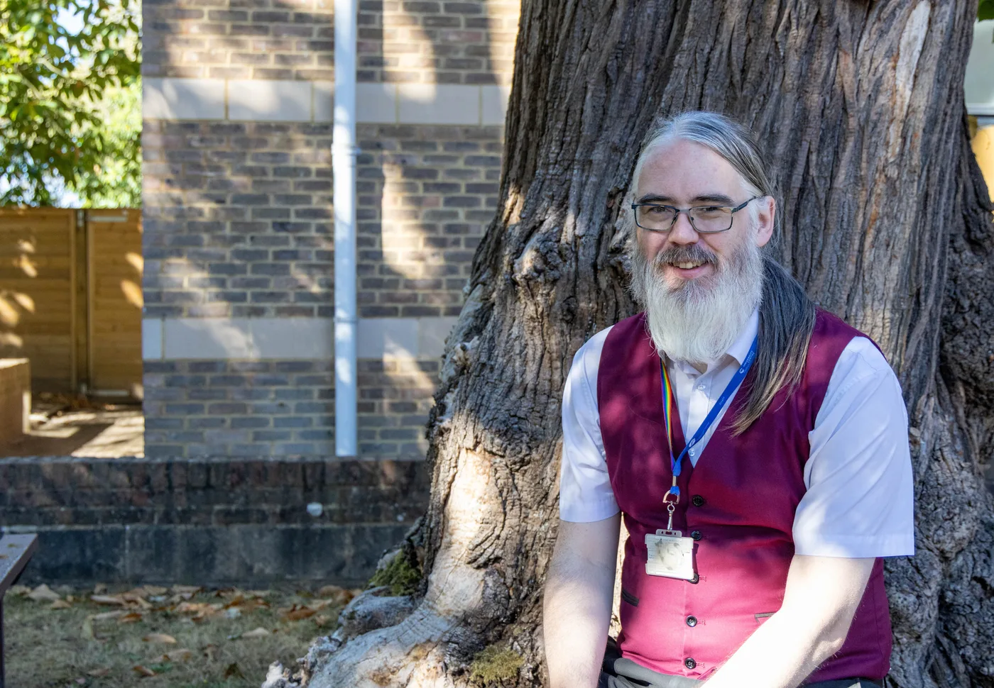Andrew Lucas, with a long grey beard, glasses, a burgundy waistcoat and lanyard, sitting in front of a large tree trunk